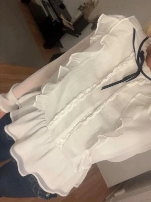 Vit volangblus med knytband - Säljer en romantisk vit blus med volangdetaljer och spets längs framsidan. Blusen har långärmad design i ett tunt, lätt material och ett svart knytband vid halsen som ger en söt kontrast. Perfekt att styla med jeans eller kjol.