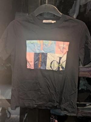 Svart Calvin Klein t-shirt med färgglad print - Svart t-shirt från Calvin Klein Jeans med en stor färgglad grafisk print framtill i blått, rosa, vitt och gult. Klassisk rund halsringning och korta ärmar. Inga synliga fläckar eller defekter. Skapa en bundle för bättre pris