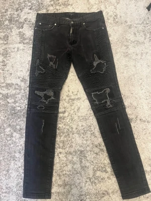 Svarta bikerjeans med slitningar - Säljer ett par svarta skinny bikerjeans med kraftiga slitningar och ribbade detaljer över knäna. Jeansen har fem fickor och dragkedjegylf. Materialet är stretchigt denim som sitter tight och ger en cool streetstil.