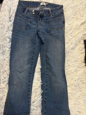 Blå bootcut paris jeans från 157 - Snygga blå bootcut jeans från 157 med två stora framfickor och dubbla knappar i midjan. Jeansen har en klassisk tvättad look och är tillverkade i denim med gul kontrastsöm. Perfekta för en avslappnad och trendig stil. Modellen heter paris