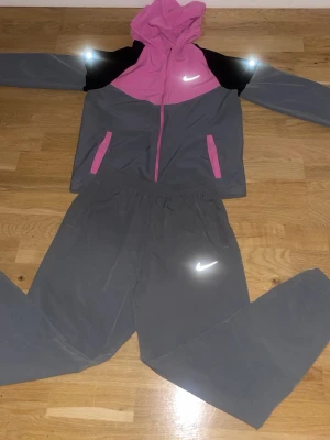 Nike grå och rosa Miler träningsset - Säljer ett snyggt träningsset från Nike med grå byxor och matchande jacka i grått, rosa och svart. Jackan har huva, dragkedja och två fickor med rosa detaljer. Både jacka och byxor har reflekterande Nike-logga. Perfekt för träning eller chill.