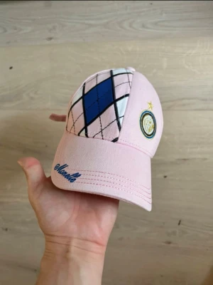  Inter Milan Rosa keps med argyle-mönster -  Inter Milan Snygg rosa keps med argyle-mönster i blått, svart och vitt framtill. Broderad logga på sidan och texten 'Monello' i blått på skärmen. Justerbar rem bak för perfekt passform. Perfekt accessoar för en trendig och sportig look.
