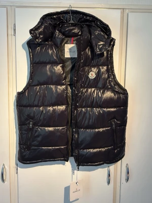 Svart dunväst från Moncler med huva - Snygg svart dunväst från Moncler med glansig finish och avtagbar huva. Västen har två dragkedjefickor fram, Moncler-logga på bröstet och quiltad design. Perfekt för lager-på-lager och håller dig varm med stil. Storlek L passar båda M och L. 