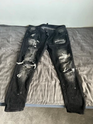 Svarta jeans dsquard2 jeans  - Coola svarta jeans med slitningar, hål och vita färgstänk. Jeansen har en smal passform och detaljer som lagningar och patchar på framsidan. Perfekta för en edgy streetstyle-look. Dsqaurd2 svarta jeans. Äkta