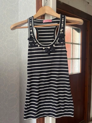 Randig linnetopp med pärlor och rosetter - Snygg svartvit randig linnetopp med ribbad struktur. Dekorerad med pärlor, små rosetter och glittriga detaljer längs ringningen. Perfekt för dig som vill sticka ut lite extra i sommarvärmen.