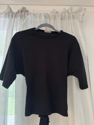 Svart knyttröja från Nelly - En svart topp med rund hals och knytband i ryggen. Toppen har korta ärmar och är gjord i ett mjukt bomullsliknande material. Perfekt för dig som gillar en enkel men trendig look. Fint skick