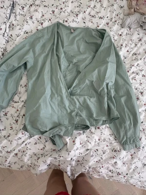 Ljusgrön omlottblus från PIECES - Snygg ljusgrön omlottblus från PIECES med långa ärmar och knytband i midjan. Blusen har en avslappnad passform och är tillverkad i ett tunt, mjukt tyg som känns skönt mot huden. Perfekt att styla med jeans eller kjol för en trendig look.