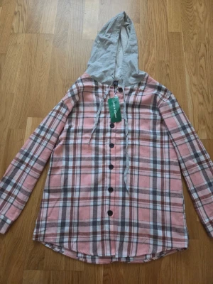 Rutig overshirt med grå huva - Säljer en snygg overshirt i rosa, grått och vitt rutmönster med långa ärmar och knäppning framtill. Skjortan har en kontrasterande grå huva med dragsko som ger en avslappnad streetkänsla. Perfekt att slänga över en t-shirt för en cool look.