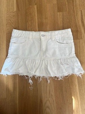 Vit volangkjol med fransig kant - Kort vit kjol i bomull med volang och fransig, rå kant nertill. Kjolen har fickor fram och bälteshällor i midjan. Perfekt för en avslappnad och trendig look.