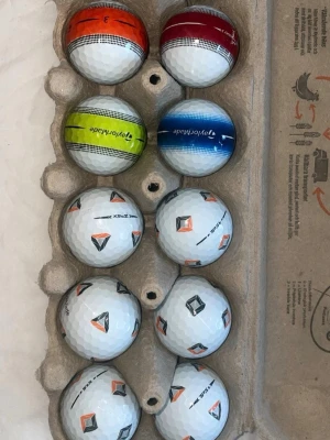 Premium bollar - Säljer ett set med 12 golfbollar i olika färger och mönster. Sex bollar är färgade i orange, röd, grön och blå med TaylorMade-tryck, medan sex bollar är vita med svart och orange logga. Perfekt för dig som vill ha variation på golfbanan.