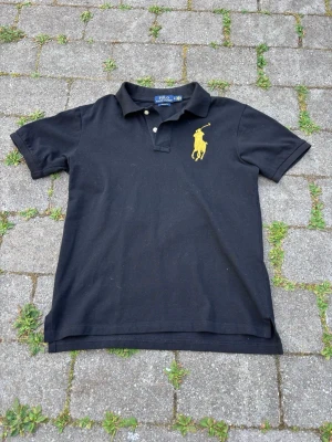 Svart pikétröja från Polo Ralph lauren - Svart pikétröja i bomull med klassisk krage och knappar framtill. Stor gul broderad logga på bröstet och en gul broderad siffra 3 på ärmen. Perfekt för en sportig och stilren look.