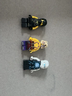 Tre LEGO-minifigurer ninja - Säljer tre LEGO-minifigurer med ninja-tema. En svart och gul figur med mask och tryck, en gul och lila figur med bar överkropp och tatueringar samt en vit och blå figur med huva och detaljerat tryck. Alla är tillverkade i plast och har rörliga armar och ben.