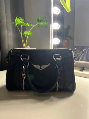 Svart handväska med silverdetaljer - Snygg svart handväska i skinnimitation med två korta handtag och silverfärgade dragkedjor framtill. Väskan har en glittrig vingdetalj på framsidan och metallringar som fästen. Perfekt för dig som gillar en edgy och stilren look. Jag köpte den i Grekland så minns inte vad butiken hette men de va en väskbutik💕