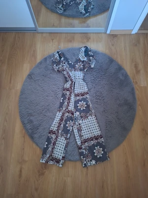 Mönstrad jumpsuit från River Island - Säljer en snygg jumpsuit från River Island med vida ben och volangdetaljer vid axlarna. Färgen är en mix av blått, beige och vinrött i ett bohemiskt mönster. Perfekt för dig som gillar statement-plagg och vill sticka ut lite extra.