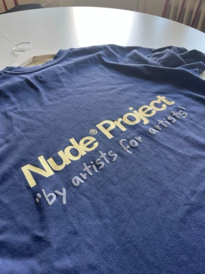 Blå Nude Project t-shirt - Snygg marinblå t-shirt från Nude Project med stort tryck på ryggen i vitt och gult. Oversized passform och klassisk rund hals. Perfekt för dig som gillar streetwear och vill sticka ut med ett coolt statement-plagg. Passar även dig med storlek M. 