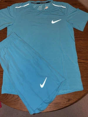 Blått träningsset Nike miler - Säljer ett blått träningsset från Nike med t-shirt och shorts. Setet har vita Nike-loggor på både tröjan och shortsen samt reflekterande detaljer på axlarna. Materialet är lätt och andas, perfekt för träning eller löpning.