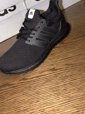 Svarta Adidas Ultraboost sneakers - Snygga helsvarta Adidas Ultraboost sneakers med stickad ovandel i mesh och klassiska tre ränder på sidan. Skorna har en bekväm och responsiv sula samt svart snörning. Perfekta för dig som gillar stilrena och sportiga sneakers.