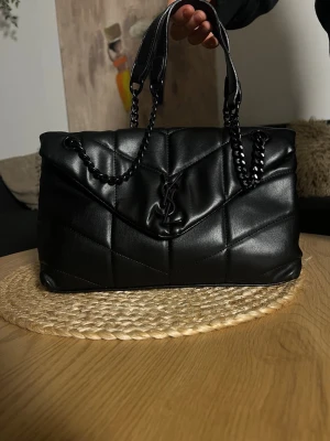 Svart quiltad handväska från YSL - Stilren svart quiltad handväska från YSL med kedjedetaljer på handtagen och YSL-logga framtill. Väskan har en mjuk, vadderad look och är tillverkad i glansigt skinnliknande material. Perfekt för dig som gillar lyxig och trendig design.
