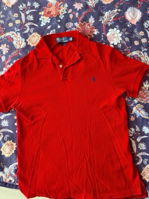 Röd pikétröja från Polo Ralph Lauren - Klassisk röd pikétröja från Polo Ralph Lauren i slim fit-modell. Tröjan har korta ärmar, krage med två knappar och den ikoniska broderade loggan på bröstet. Perfekt för en stilren och avslappnad look.