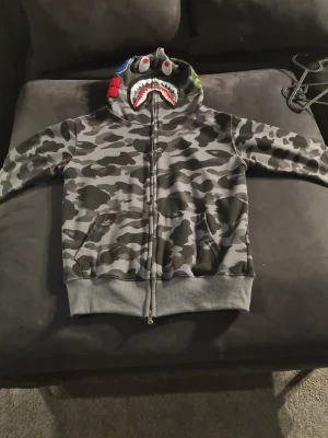 Grå camo hoodie med hajmotiv - Säljer en grå camouflagemönstrad hoodie med dragkedja och huva. Huvan har ett hajansikte med tänder och ögon, samt färgglada detaljer på sidorna. Tröjan har ribbade muddar och fickor framtill. Perfekt för dig som vill sticka ut med streetwear-stil.