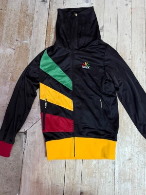 Svart hoodie med färgglada detaljer - Säljer en svart hoodie från Skank med dragkedja och huva. Framsidan har diagonala ränder i grönt, gult och rött samt gul mudd nedtill och en röd och grön mudd vid ärmsluten. Två fickor med dragkedja och broderad logga på bröstet. Perfekt för dig som gillar streetwear och färg.