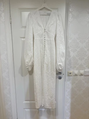 Helt ny vit långklänning från Adoore - Elegant vit långklänning från Adoore i blommigt mönstrat tyg. Klänningen har v-ringning, långa puffärmar och knappar framtill. Materialet har en lätt glans och broderade detaljer som ger en exklusiv känsla. Perfekt för speciella tillfällen.
