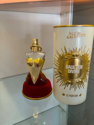 Gaultier Divine Le Parfum - Unik parfymflaska från Jean Paul Gaultier formad som en kvinnokropp med guldfärgade detaljer och lock. Flaskan står på en röd sammetsbas och kommer med en elegant vit och guldig förpackning med solstråle-design. Perfekt för dig som gillar lyxiga och iögonfallande dofter.