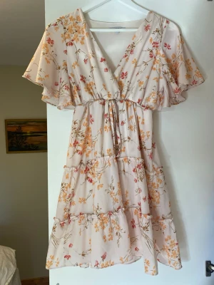 Blommig kortklänning med volang - Söt kortklänning i ljusbeige med blommigt mönster i gult, orange och rosa. Klänningen har v-ringning, volangärmar och omlottkänsla framtill samt knyt i midjan. Perfekt för sommarens varma dagar.