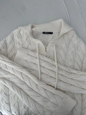 Vit kabelstickad hoodie Gina Tricot - Säljer en krämvit kabelstickad hoodie från Gina Tricot. Tröjan har en mysig huva med dragsko, ribbade muddar och ett snyggt flätmönster över hela plagget. Perfekt för dig som gillar en chill och trendig stil.