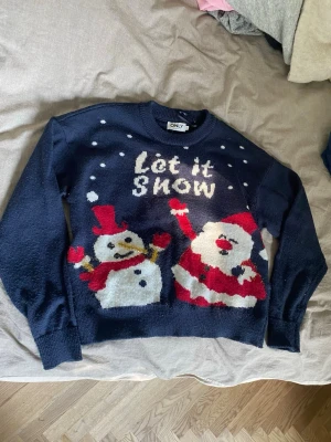Mörkblå jultröja med snögubbe & tomte - Mysig mörkblå stickad jultröja från ONLY med motiv av snögubbe och tomte samt texten 'Let it Snow' framtill. Tröjan har långa ärmar och ribbade muddar vid ärmslut och nederkant. Perfekt för vinterns alla julmys!  Använd endast 2 gånger 