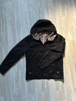 Svart vindjacka med rutigt foder - Snygg svart vindjacka med huva och dragkedja. Insidan av huvan har ett klassiskt beige, rött och svart rutigt mönster. Jackan har två fickor med dragkedja och diskret broderad logga på bröstet. Perfekt för blåsiga dagar.