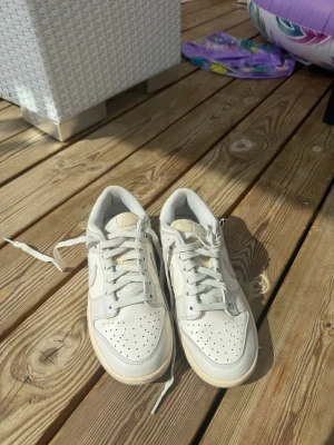 Low Nike Dunks - Snygga vita sneakers i lågt modell med beige sula och klassisk snörning. Skorna har perforerad tå för extra ventilation och en stilren design som passar till det mesta. Tillverkade i läder med textilfoder för skön komfort.