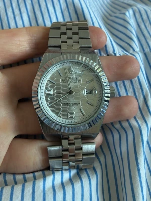 Rolx  - Stilren klocka från Rolx i silverfärgat metall med klassisk länkarmband och räfflad bezel. Urtavlan har ett vågigt mönster och tydliga siffror vid 6 och 9 samt datumfönster. Perfekt accessoar för dig som gillar exklusiv stil. Säljer billigt pågrund av spricka i glaset 