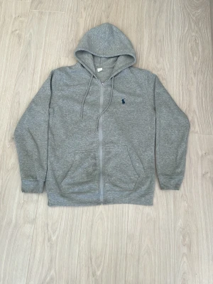 Polo Ralph Lauren Hoodie - Snygg grå hoodie med dragkedja från Polo Ralph Lauren. Klassisk design med huva och dragsnören, två fickor framtill och den ikoniska blå broderade loggan på bröstet. Tillverkad i mjukt sweatshirtmaterial som är perfekt för chill dagar.
