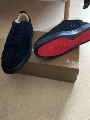 Svarta sneakers med nitar och röd sula - Snygga svarta sneakers från Christian Louboutin med ikonisk röd sula och svarta nitar på tån. Skorna har en låg profil, svarta skosnören och är tillverkade i mocka och läder. Perfekta för dig som vill sticka ut med en edgy och exklusiv stil.