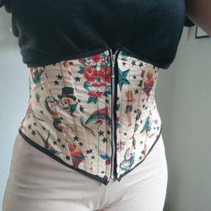 Korsett stålskena underbust pinup tatuering - Liten rolig korsett med fint traditionellt tatueringsmönster som fungerar perfekt som dekorationsbälte eller för att ge litet form. Dragkedja fram, liten söm som gått (se bild). Tunna stålskenor, kan reducera, men är mer dekorativ än den kan komprimera. 