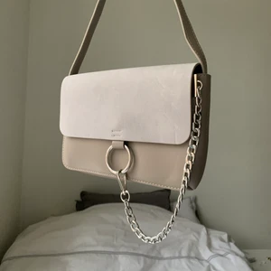 Axelremsväska!  - Grå/beige axelremsväska från NA-KD i PU skinn med locket i mockaimitation. Använd vid något enstaka tillfälle. Tyvärr lite skadad under till (se bild 3), i övrigt är den i mycket bra skick!