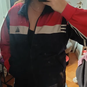 Adidas Övergångsjacka - SKITSNYGG Adidas Övergångsjacka i 100% polyester med rött, blått och vitt med svarta detaljer. Perfekt för idrottslektionerna eller om man behöver ett lätt plagg när det blåser lite ute, supertrendig och går att använda när som helst! Har fickor med dragkedja. Användes senast för 3 år sedan men använder den inte längre! Lite små trådar syns från ryggloggan på insidan och lite nopprig på insidan längst resoren längst ner. Köpt på HUMANA för 349kr. 