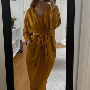 Senapsgul långklänning - Fin långklänning med knyte där i midjan. Man kan välja vart man vill att rosetten ska sitta. Slits framtill, vilket gör det lättare att gå. Använd 1 gång! Köpt för 599 200kr