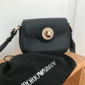 ÄKTA emporio armani väska - Säljer nu denna fina emporio armani väska, använd endast vid två tillfällen och då i bara några få timmar. Axelbandet är långt och brett och är därför mycket bekväm att bära❤️ dustbag ingår. Den har en PYTTELITEN skrapa som jag märkte när jag tog bilder på ”knappen” men absolut inget man tänker på. Över lag i mycket gott skick! Köparen står för frakten/det fraktalternativ som föredras🥰