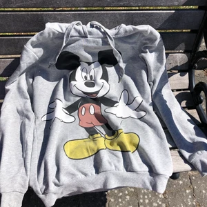 Grå hoodie i storlek M. - Grå hoodie med Musse Pigg-tryck. Materialet är något tunnare än på en ”vanlig” hoodie så den är inte lika varm. Har luva med snören, resår i midjan och armarna men saknar fick.