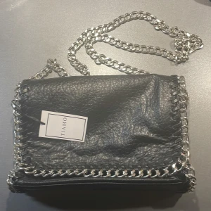 Oanvänd svart handväska  - Svart väska med silverkedja, lappen sitter kvar, helt oanvänd, märket ”tiamo” 