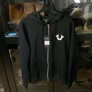 True Religion Zip up L (liten i storlek)  - True Religion zip up huvtröja.      Äkta (QR kod finns).  Sugg retail price 1265:- ekl. Moms. Perfekt skick helt ny aldrig använd.  Köpte för 1582 kr inkl. Moms och tullavgift från Miami, USA.  Storlek L men liten i storlek.  Pris kan diskuteras. 