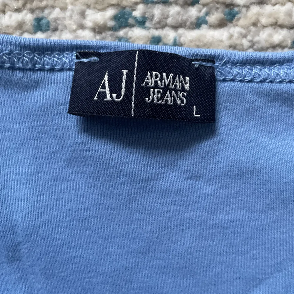 en ljusblå t-shirt från märket armani jeans som jag köpt second hand! dragkedjan är justerbar och det sitter rhinestones/strass vid kragen🥰skulle säga att den passar en storlek M också då den är liten i storleken🫶(jag vet inte om den är äkta!!). T-shirts.