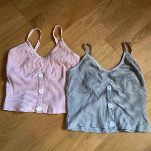 2 Crop-top Linnen - 2 croppade och ribbade linnen med knapp-detalj frampå. Den ena är en ljus rosa och den andra en ljus grå-grön.   Dom är i jättefint skick, inte noppiga eller trasiga på något vis. Säljer då jag växt ur dom.
