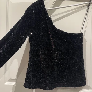 Svart Glittrig One shoulder - Säljer nu den perfekta party toppen, sprillans ny med taggar kvar. Storlek M. Köpt från Bikbok för 299kr säljs nu för 170kr. Fraktas på köparens bekostnad