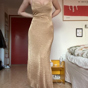 Guldig långklänning - Långklänning med guldigt tyg och dubbel slits med spetsband. Spetsbanden går att ta bort. Tyget har ett fåtal skador där trådar dragits ut lite men inget som märks. Passar någon som kan bära utan bh då ryggen är lite låg. Jag är 173cm, bär S-M, Bh 70F