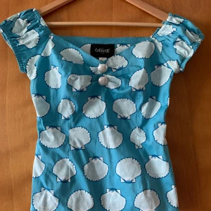 Collectif retro topp snäckor - Oanvänd topp från Collectif. Snäckmönster i vitt mot ljusblå bakgrund och pärlemorknappar framtill. Storlek S