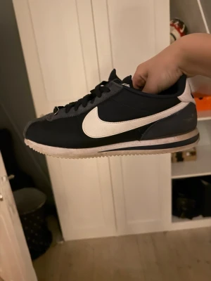 Nike Cortez svartvita sneakers - Säljer ett par klassiska Nike Cortez sneakers i svart med vit sula och vit swoosh på sidan. Skorna har låga skaft, snörning och yttermaterial i textil och syntet. Sulan är räfflad för bra grepp och retrovibbarna är på topp.