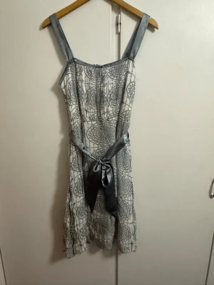 Vit och grå kortklänning med mönster från Flash - Snygg kortklänning i vitt med grått grafiskt mönster och breda axelband. Klänningen har ett matchande grått band i midjan som knyts till rosett och ger en feminin siluett. Perfekt för sommarens alla tillfällen.